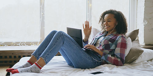 Young woman in jeans and checkered shirt waving to therapist online using her laptop