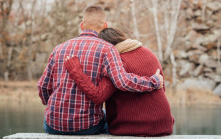 Image of a couple supporting each other sitting on a stone bench and looking at a pond.